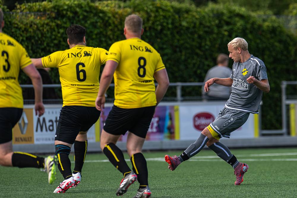 Drie spelers zetten aan; de bal rolt tussen geel en grijs door.
