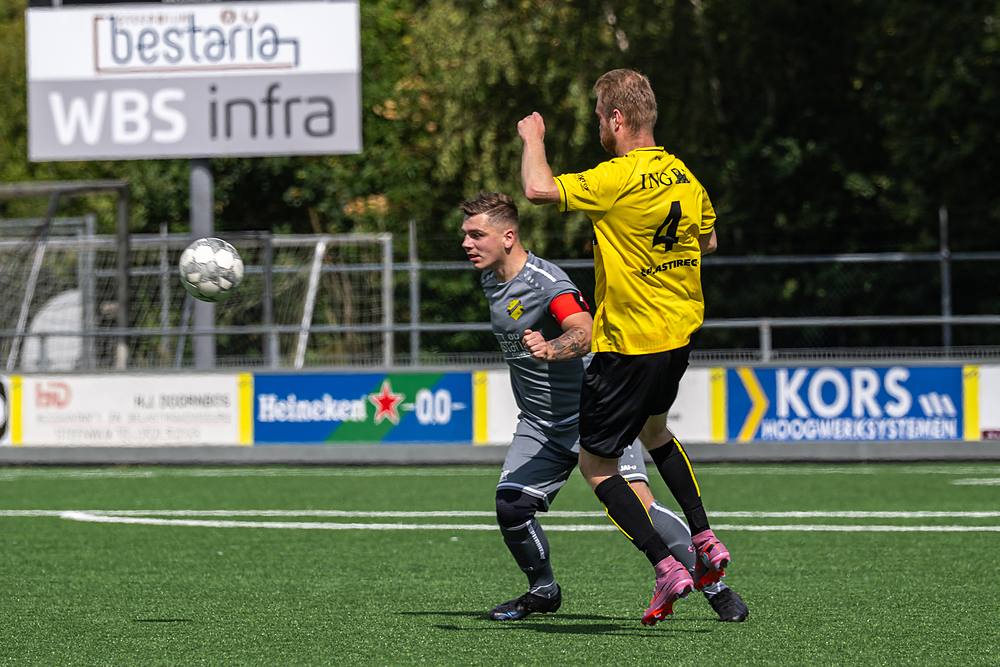 Speler in het geel gaat het luchtduel aan met een tegenstander in het grijs.