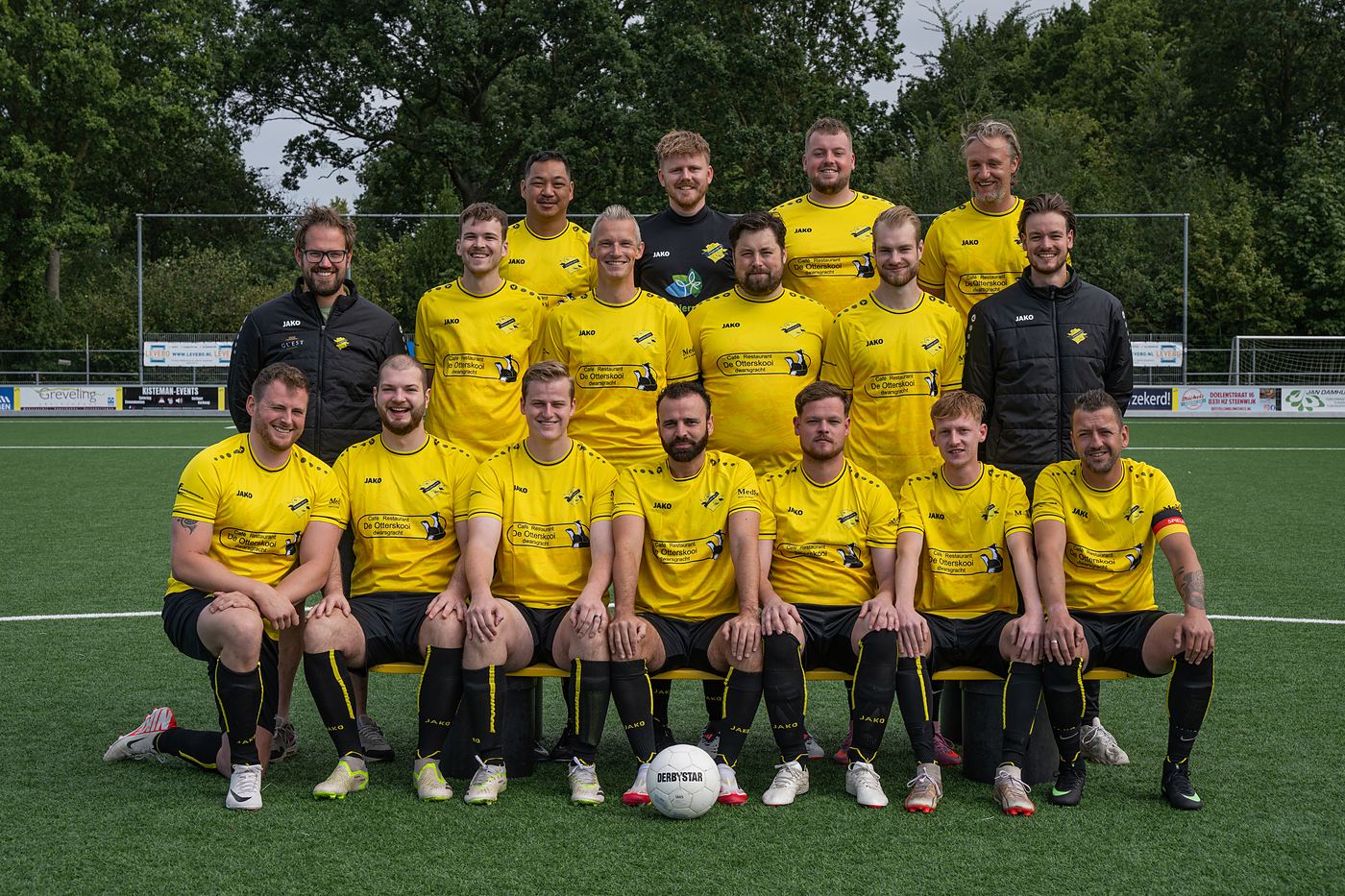 Teamfoto van Zondag 2 van vv Steenwijker Boys op het kunstgras: veertien spelers, een keeper in het zwart en twee leiders, in het gele thuisshirt met Caf&eacute; Restaurant De Otterskooi op de borst.